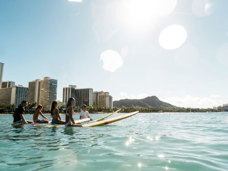 People paddleboarding together on turquoise water near a city skyline with high-rise buildings and a distant hill under a sunny sky.