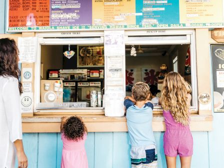 Seaside food stand with colorful menu boards, a window service, and kids ordering at the counter under a corrugated roof.