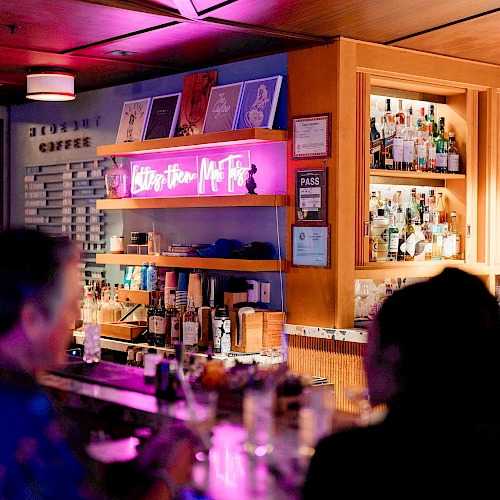 Bar scene with warm lighting, shelves of bottles, neon sign, and two patrons facing the bar.