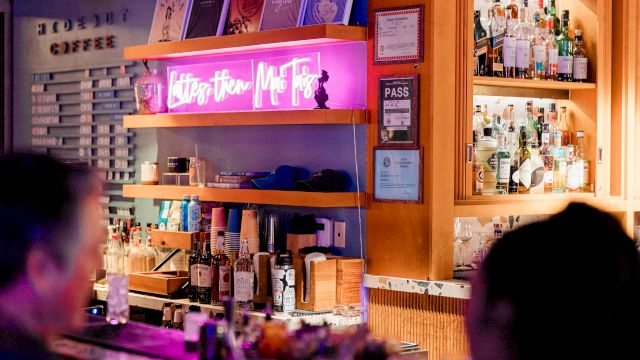 Bar scene with warm lighting, shelves of bottles, neon sign, and two patrons facing the bar.