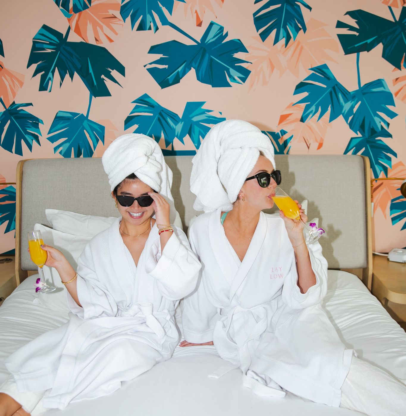 Two people in white bathrobes with towels on their heads sit on a bed, sipping drinks, wearing sunglasses, in a tropical leaf wallpaper room.
