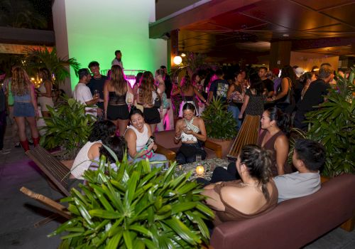 A crowded party at night with people mingling on couches and around plants, neon lighting, and a lively social vibe, everyone enjoying themselves.
