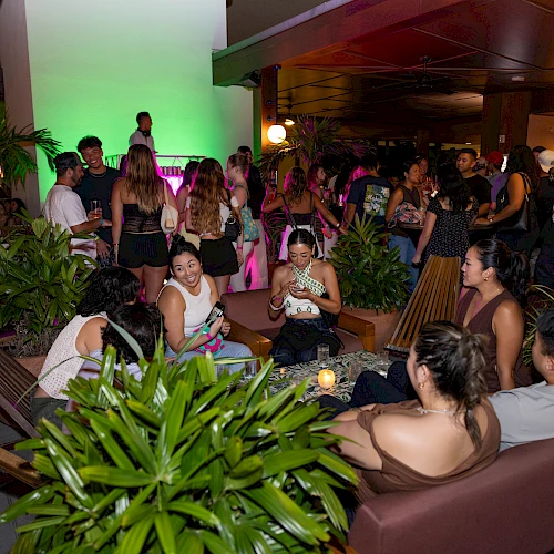 A crowded party at night with people mingling on couches and around plants, neon lighting, and a lively social vibe, everyone enjoying themselves.