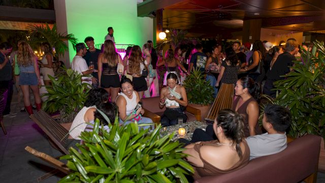 A crowded party at night with people mingling on couches and around plants, neon lighting, and a lively social vibe, everyone enjoying themselves.