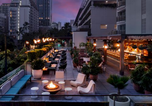 A city rooftop lounge at dusk with palm plants, outdoor seating, warm lights, and fire pits overlooking tall buildings.
