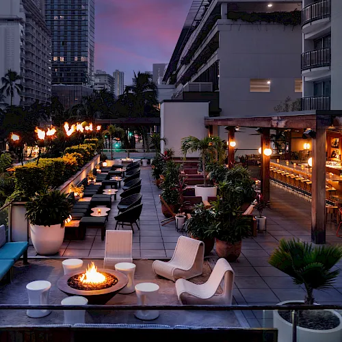 A city rooftop lounge at dusk with palm plants, outdoor seating, warm lights, and fire pits overlooking tall buildings.