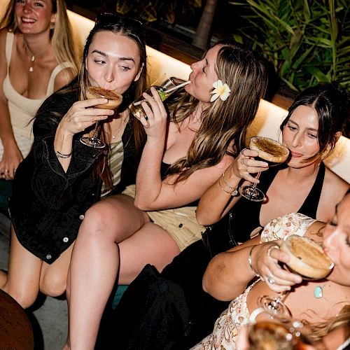 A group of stylish women are lounging and toasting with cocktails at a nighttime outdoor party, smiling and posing for a selfie.