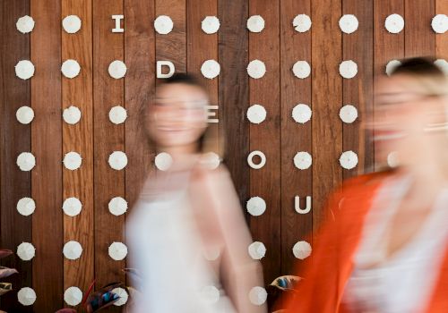 A rustic wooden wall covered with white pushpins and letter tiles as people walk by.