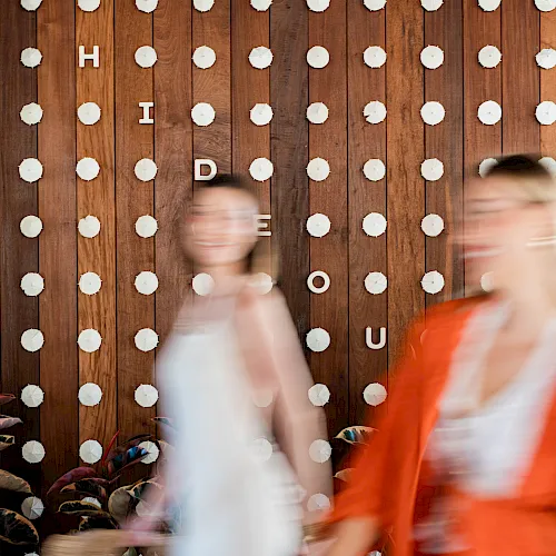 A rustic wooden wall covered with white pushpins and letter tiles as people walk by.