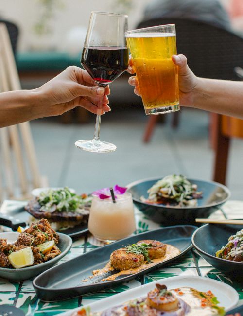 Two people clink glasses of dark red wine and golden beer over a table of food at an outdoor meal.