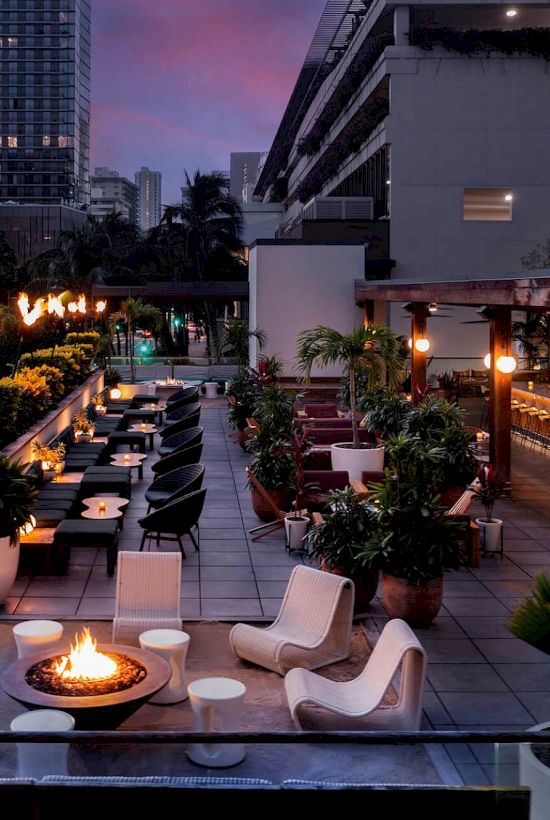 A rooftop hotel terrace at dusk with potted plants, lounge seating, a fire pit, and a lit bar, surrounded by tall city buildings.