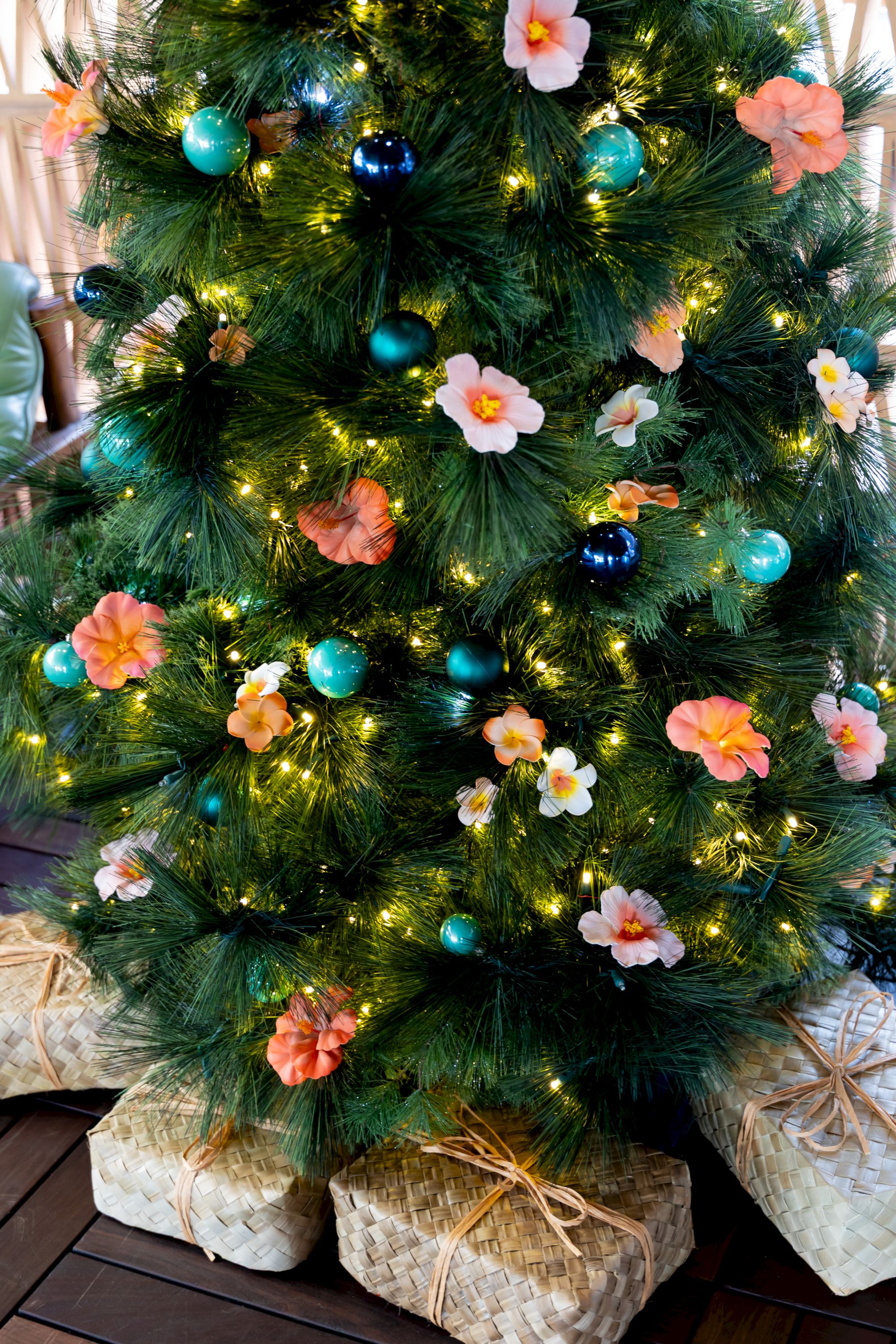 A decorated Christmas tree with teal and blue baubles, glowing lights, and pastel flowers, set on woven wicker gifts.