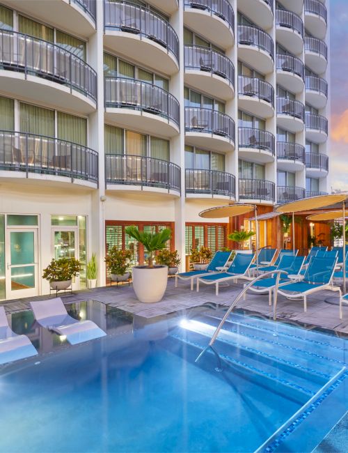 A hotel pool terrace with blue loungers, umbrellas, plants, and a multi-story balcony building under a colorful sky.