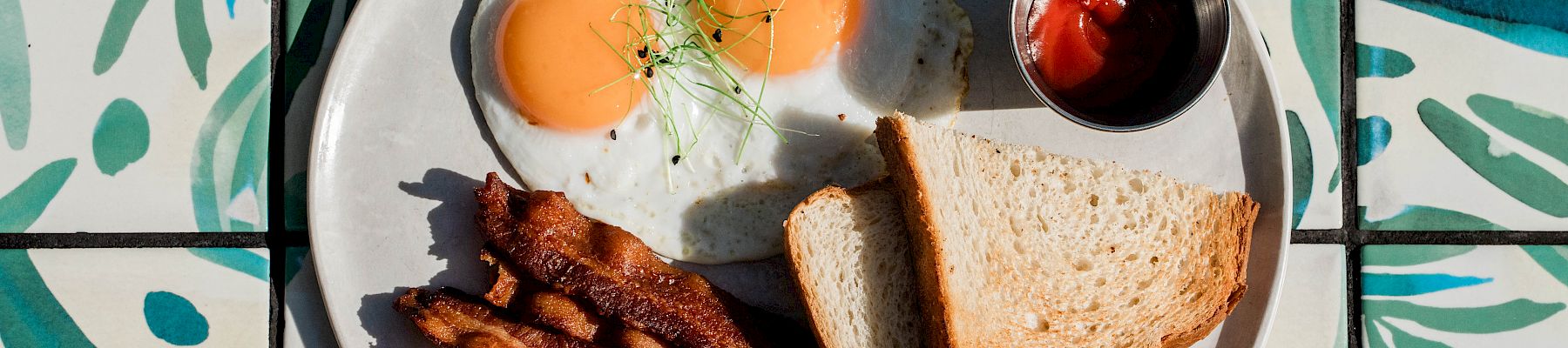 A breakfast plate with sunny-side eggs, bacon strips, toast, and a small cup of ketchup/berries, on a patterned tile background.