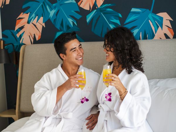 A couple in white robes clink orange drinks on a sofa, smiling under a bold teal-and-orange wall mural.