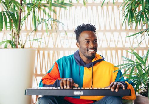 A smiling man in a colorful hoodie plays a KORO keyboard by bright windows, with green plants nearby.