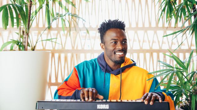 A smiling man in a colorful hoodie plays a KORO keyboard by bright windows, with green plants nearby.