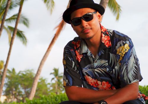 A man in a black hat and sunglasses, wearing a floral shirt, relaxing by palm trees in a tropical outdoor setting.