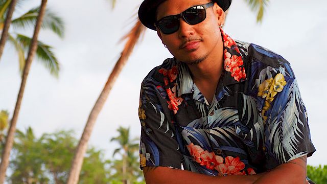 A man in a black hat and sunglasses, wearing a floral shirt, relaxing by palm trees in a tropical outdoor setting.