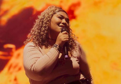 A woman with curly hair sings into a microphone on stage under warm orange concert lights, smiling as she performs.