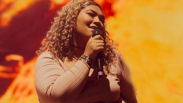 A woman with curly hair sings into a microphone on stage under warm orange concert lights, smiling as she performs.