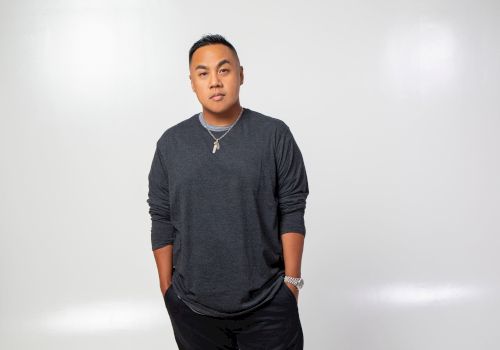 A man poses in a studio against a plain white background, wearing a dark long-sleeve shirt, necklace, and watch.