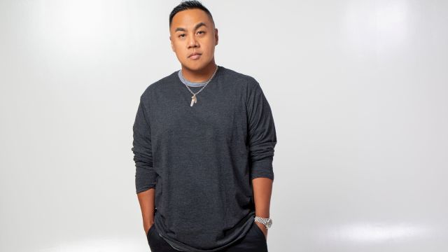 A man poses in a studio against a plain white background, wearing a dark long-sleeve shirt, necklace, and watch.