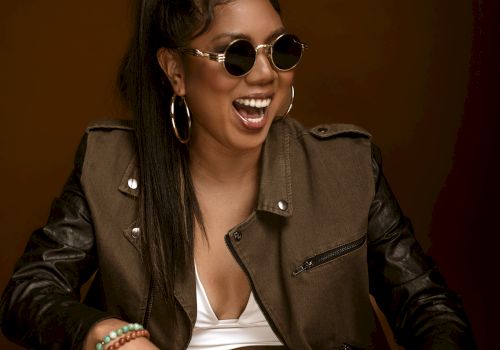 Smiling woman wearing round sunglasses and hoop earrings in a studio portrait with a leather jacket against a brown backdrop.