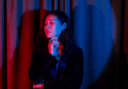 A woman in a dark suit poses with red-blue stage lighting, hand near her chin, standing before dark curtains.