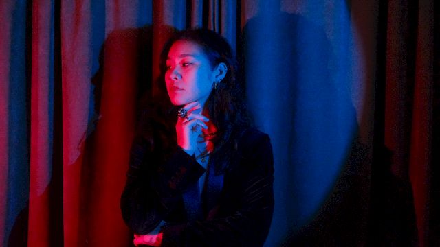 A woman in a dark suit poses with red-blue stage lighting, hand near her chin, standing before dark curtains.