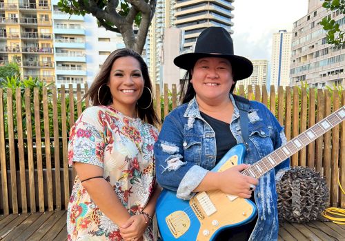 Two smiling women pose outdoors; one holds a blue electric guitar on a patio with city apartments behind them.