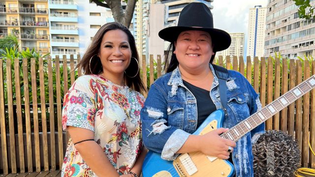 Two smiling women pose outdoors; one holds a blue electric guitar on a patio with city apartments behind them.