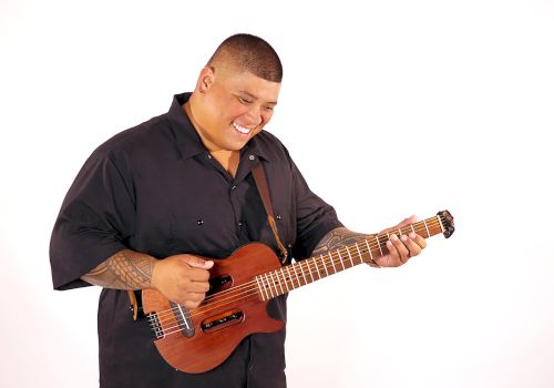 A smiling man plays a brown electric guitar, leaning forward with his fingers on the frets.