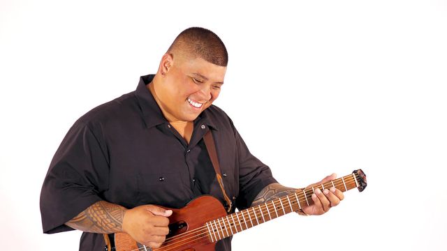 A smiling man plays a brown electric guitar, leaning forward with his fingers on the frets.
