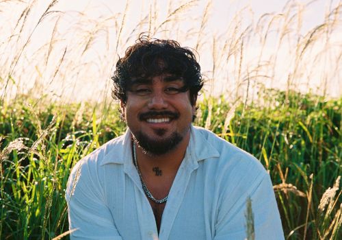A smiling man sits in a sunlit field of tall grass, looking at the camera.