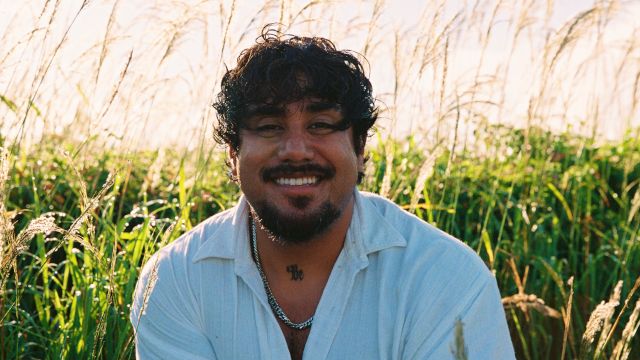 A smiling man sits in a sunlit field of tall grass, looking at the camera.