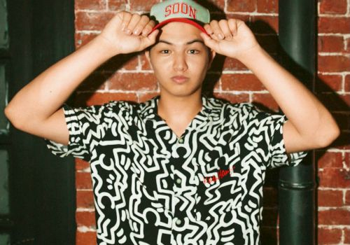 A teen in a patterned button-up and cap marked &ldquo;500N&rdquo; poses with both hands on his hat against a brick wall, relaxed.