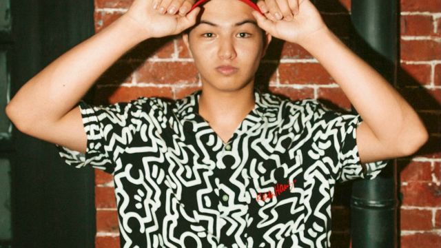 A teen in a patterned button-up and cap marked &ldquo;500N&rdquo; poses with both hands on his hat against a brick wall, relaxed.