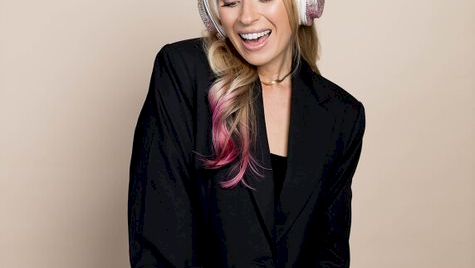 A smiling woman in a black blazer dress wearing over-ear headphones stands against a plain beige background with her hands clasped.