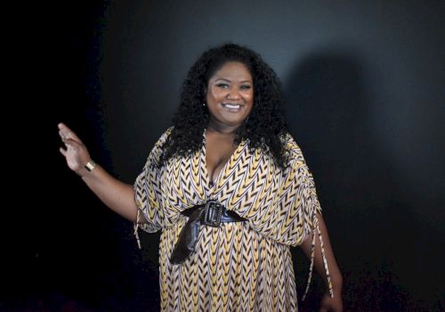 A woman with long curly hair in a patterned yellow-and-white dress smiles and gestures beside a dark studio backdrop.
