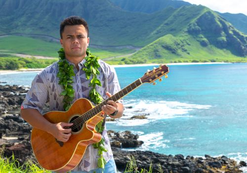 A man strums an acoustic guitar with a green lei at a rocky beach, with turquoise ocean and green mountains behind him.