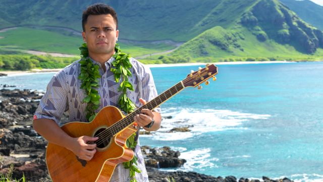 A man strums an acoustic guitar with a green lei at a rocky beach, with turquoise ocean and green mountains behind him.