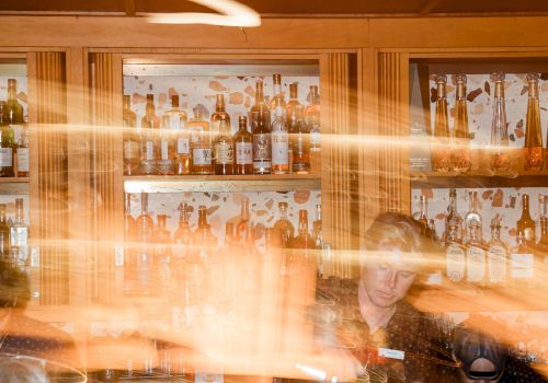 A bar liquor shelf packed with bottles, warm lights, and a bartender with motion blur in the foreground behind the glass.