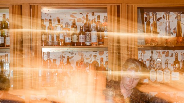 A bar liquor shelf packed with bottles, warm lights, and a bartender with motion blur in the foreground behind the glass.