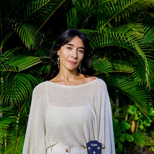 A person poses outdoors in front of lush palm leaves, wearing a light top and holding a guitar.
