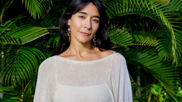 A person poses outdoors in front of lush palm leaves, wearing a light top and holding a guitar.