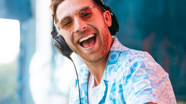 A smiling DJ wearing over-ear headphones and mirrored sunglasses, grinning into a microphone during a lively performance.