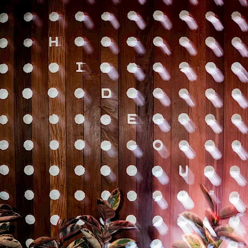 A wooden fence covered in drifting white umbrellas, with letter tiles spelling &ldquo;HIDE&rdquo; amid the scene.